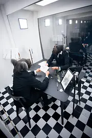 Ein Mann mit Hundemaske über dem Kopf sitzt an einem Schreibtisch, hält einen Zettel in der Hand und sieht sich im Spiegel. Schwarz-weiße Fliesen bedecken den Boden.